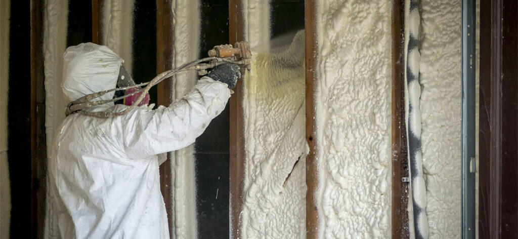 A man dressed in a white suit is spraying insulation onto a wall, demonstrating proper application techniques for insulation.